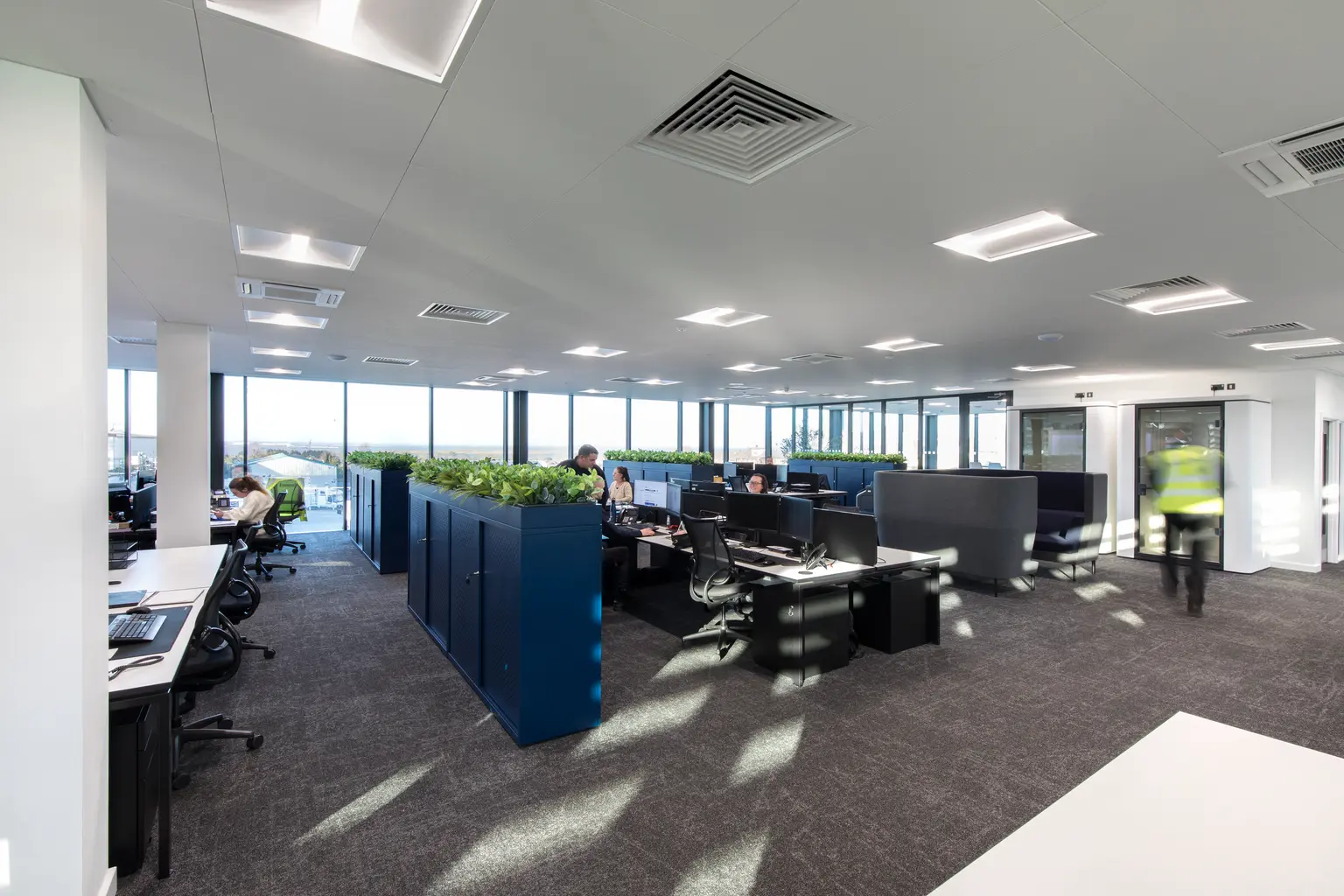 MEMS Power Generation Headquarters, Gillingham - Modern open-plan office with desk clusters arranged between navy blue storage partitions topped with greenery. Several employees are working at computer stations, while large floor-to-ceiling windows provide abundant natural light throughout the contemporary workspace