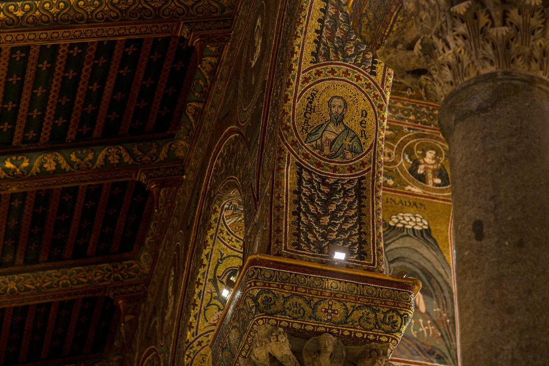 Close-up of richly decorated columns and arches with golden mosaics and depictions of saints in a church. The architecture showcases artistic ornaments, religious motifs, and an intricately crafted wooden ceiling.