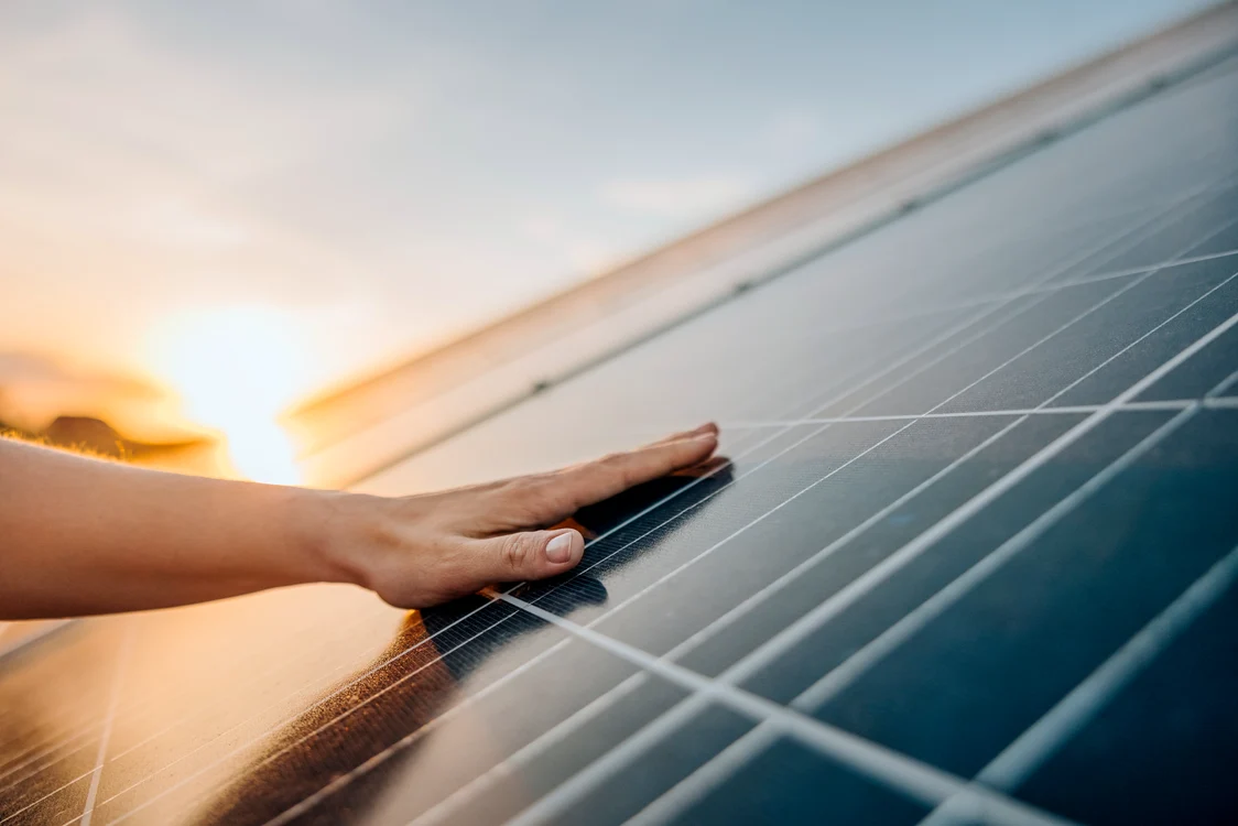 A hand touches a reflective photovoltaic surface at sunset, with sunlight reflecting off the solar modules.