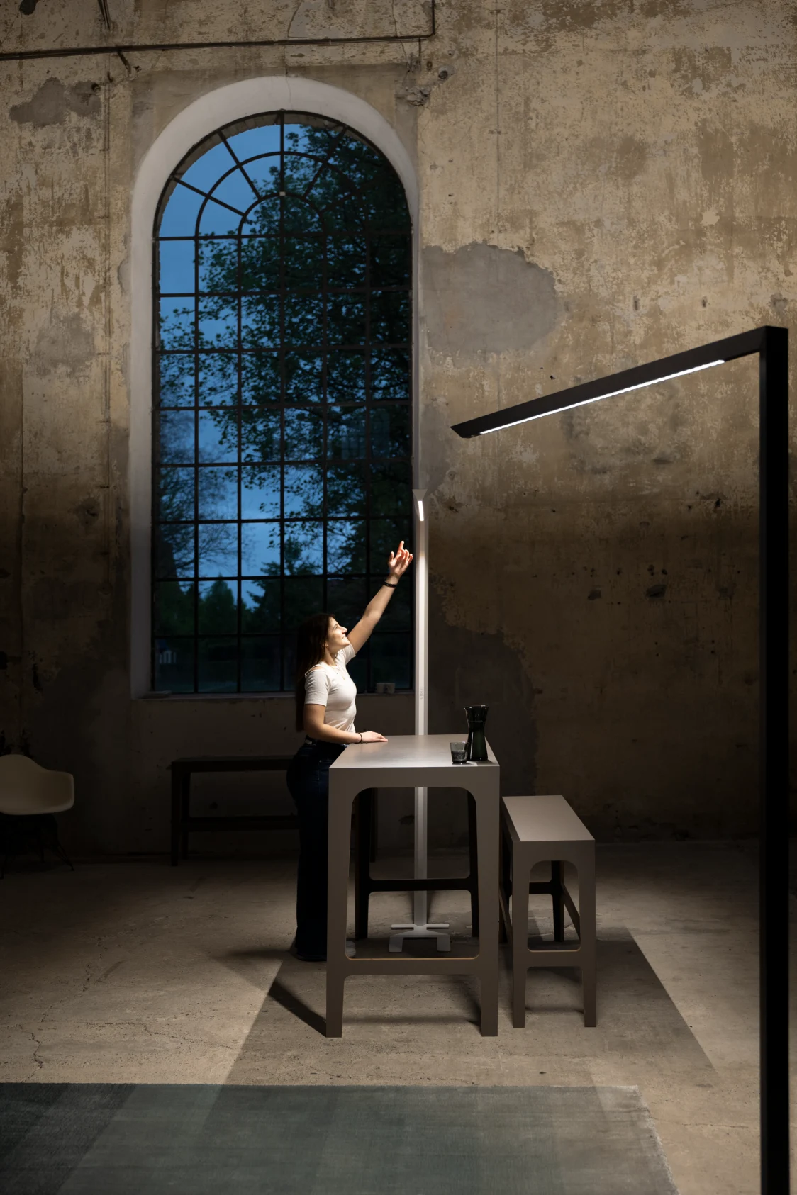 Une personne et deux lampes de table Skena dans un espace industriel haut de plafond auquel les luminaires confèrent une ambiance architecturale moderne. 