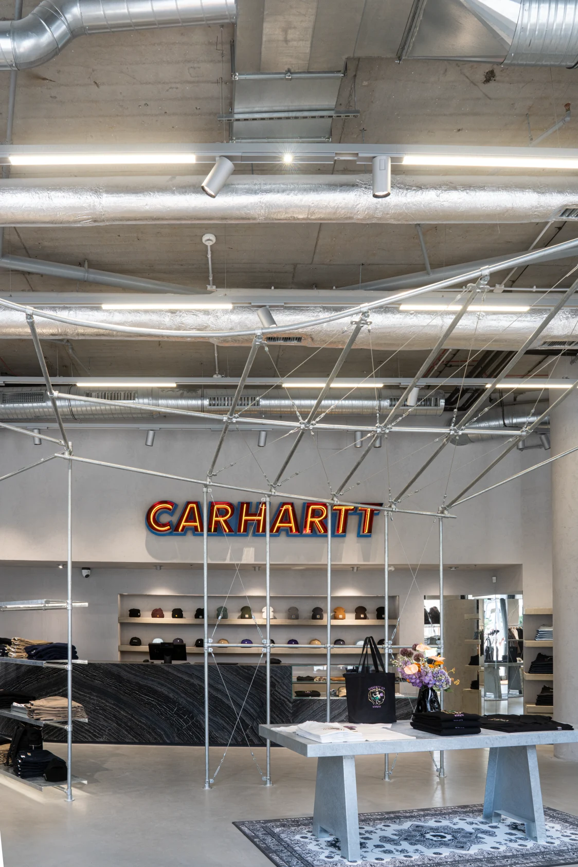 Carhartt-WIP-Store mit industriellem Design, sichtbaren Lüftungsrohren und maßgeschneidertem Lichtkonzept von Zumtobel. Im Vordergrund Verkaufstisch mit Accessoires, im Hintergrund beleuchtetes Carhartt-Logo und Regale mit Caps.