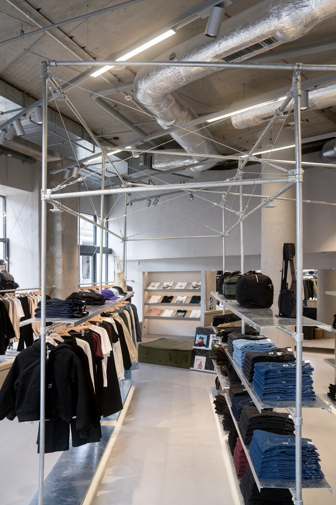 Carhartt-WIP-Store mit industriellem Design, sichtbaren Metallstrukturen und Lüftungsrohren, modernes Lichtkonzept von Zumtobel. Im Vordergrund gefaltete Jeans und Kleidung auf Kleiderstangen, im Hintergrund Regale mit Accessoires und Büchern.