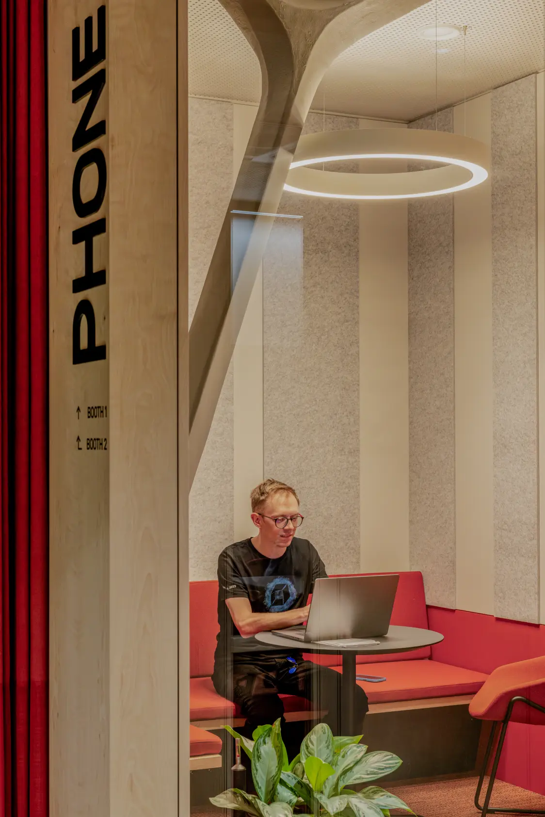 Moderne Büro-Telefonzelle mit roter Sitzgelegenheit, einem Laptop auf einem kleinen Tisch, vertikalen akustischen Wandpaneelen und einer runden Deckenleuchte.