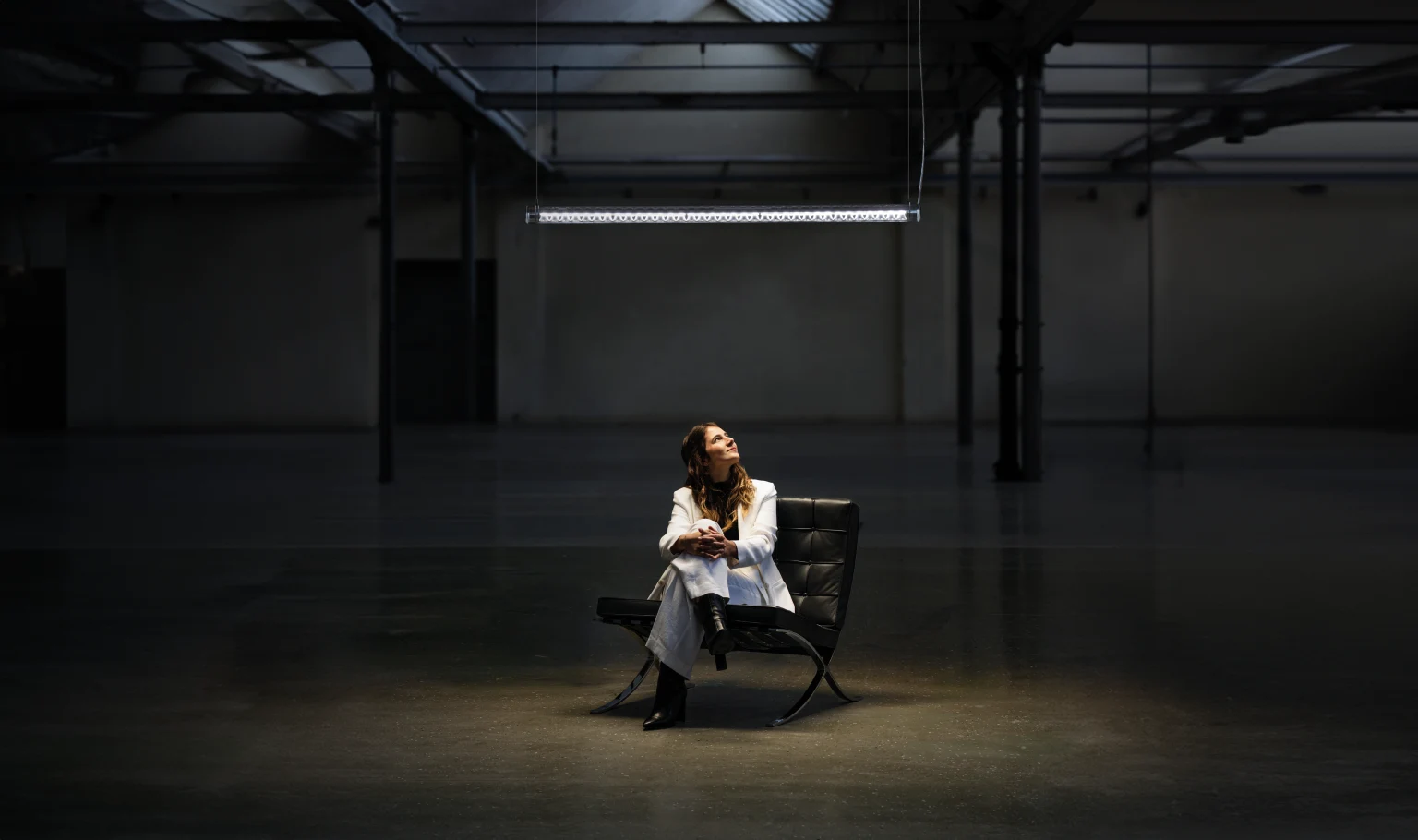 A woman sitting alone on a modern black lounge chair in a large, dimly lit industrial hall. She wears a light-colored suit and looks upward toward a single suspended linear light fixture above her. The spacious interior features high ceilings, exposed beams, and metal columns, with most of the space remaining dark except for the illuminated area around her.