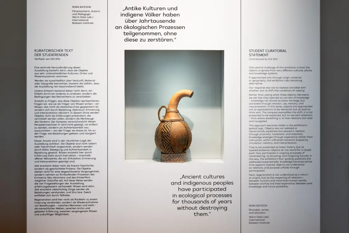 A museum display featuring a carved vessel with a curved spout positioned in a recessed wall niche. The vessel is centered between two large panels of printed exhibition text in different languages. The background panels discuss themes of ancient cultures, ecological processes, and curatorial statements.