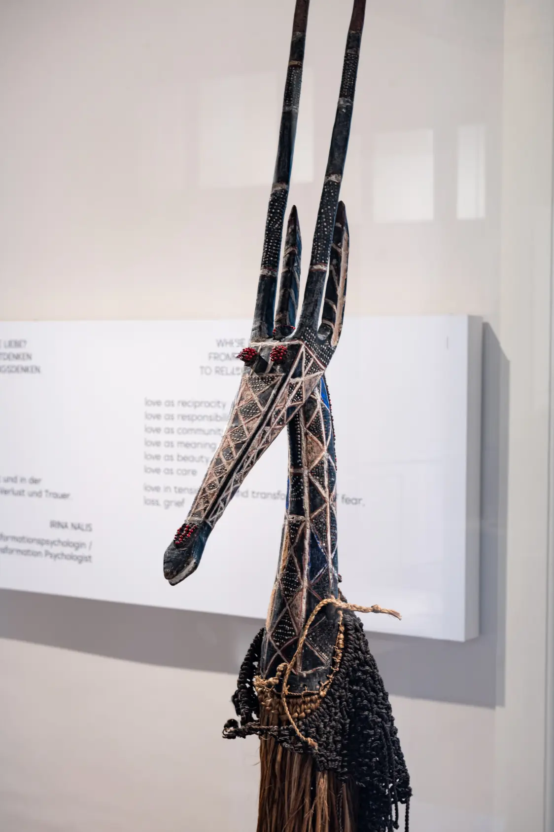 A detailed carved sculpture with an elongated, patterned head and tall vertical horns is displayed in a museum setting. The surface features geometric designs and bead embellishments. Behind the sculpture, a white exhibition panel with printed text is visible on the wall.