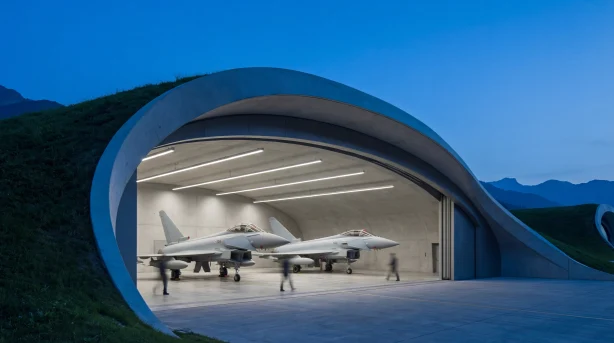 Twee moderne gevechtsvliegtuigen geparkeerd in een strakke, gebogen hangar die in een heuvel is gebouwd. De hangar heeft een futuristische, grotachtige opening en heldere binnenverlichting. Enkele mensen staan bij het vliegtuig, en bij schemering zijn bergen zichtbaar op de achtergrond.