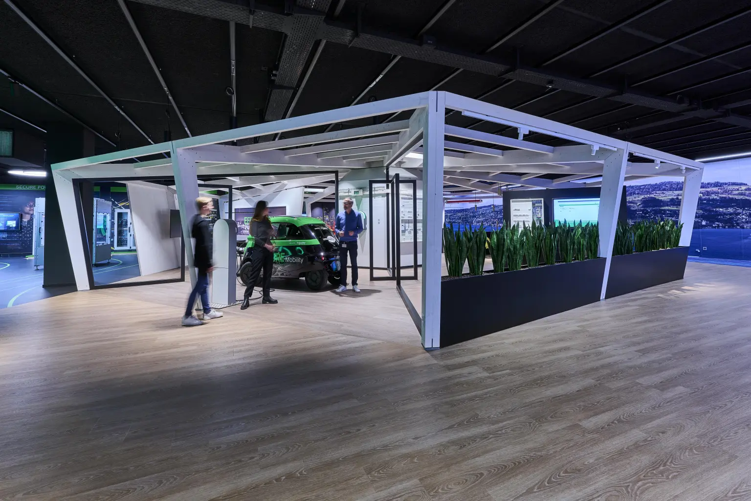 Moderner Ausstellungsbereich mit einer offenen Holzstruktur, Displays und Pflanzen, in deren Mitte ein kleines Elektrofahrzeug steht. Besucher bewegen sich durch den Raum und betrachten die gezeigten Energie- und Mobilitätslösungen.