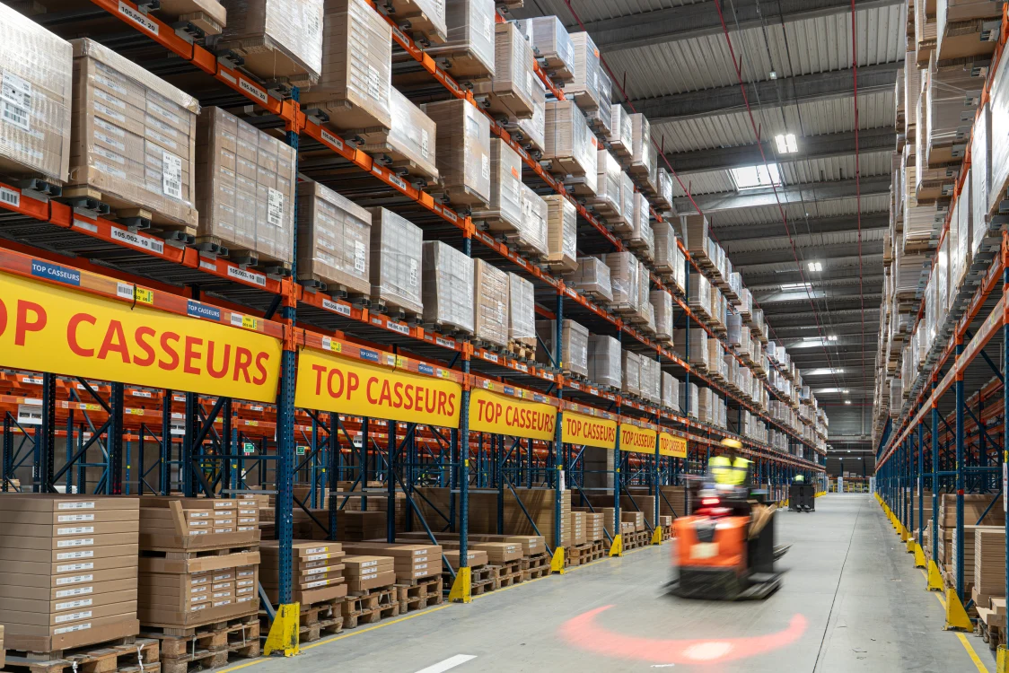IKEA warehouse interior with high-bay storage racks, palletized goods, forklift traffic, and modern industrial LED lighting.