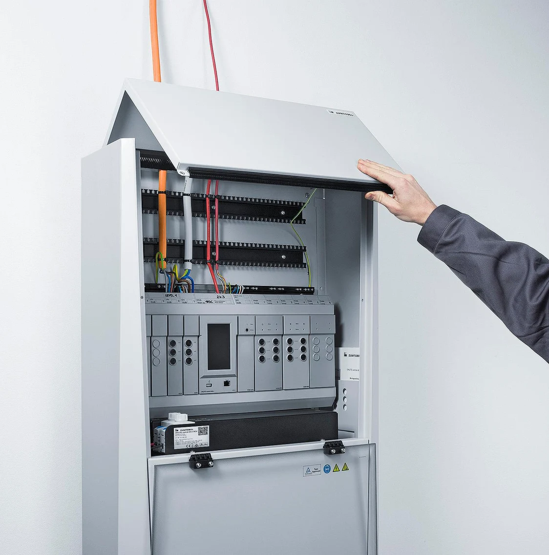  The cabinet of the ONLITE central eBox emergency power supply system is opened by a hand, revealing cables and electronic components inside.