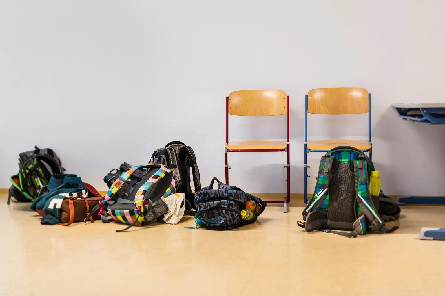 Mehrere Rucksäcke stehen auf dem Boden neben zwei Holzstühlen vor einer schlichten weißen Wand. Die Szene spielt in einem Klassenzimmer mit hellem Bodenbelag.