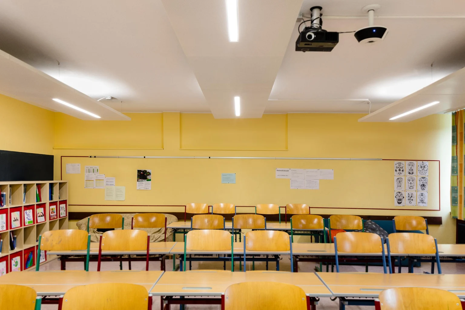 Ein helles und organisiertes Klassenzimmer mit gelben Wänden, Holzschreibtischen und passenden Stühlen. Der Raum verfügt über ein Whiteboard, Ablageflächen und an der Decke montierte Lampen für optimale Ausleuchtung.