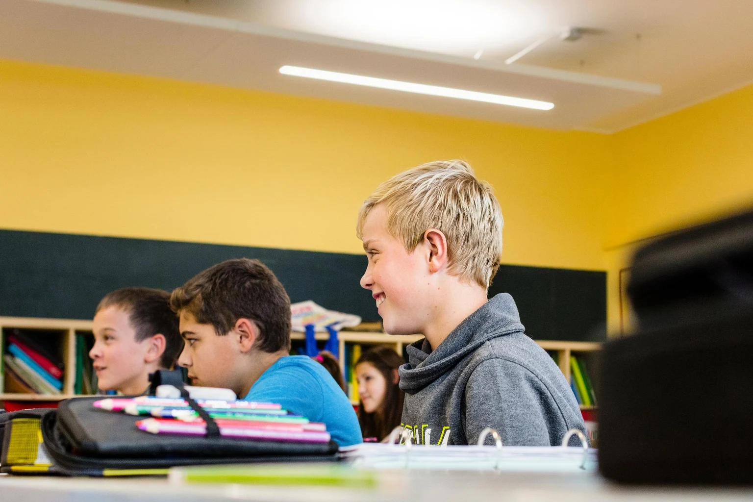 Die Schüler sitzen in einem Klassenzimmer mit gelben Wänden, einer grünen Kreidetafel und Regalen voller bunter Bücher und Materialien. Der Raum ist mit Deckenleuchten hell erleuchtet und bietet eine konzentrierte Lernatmosphäre.