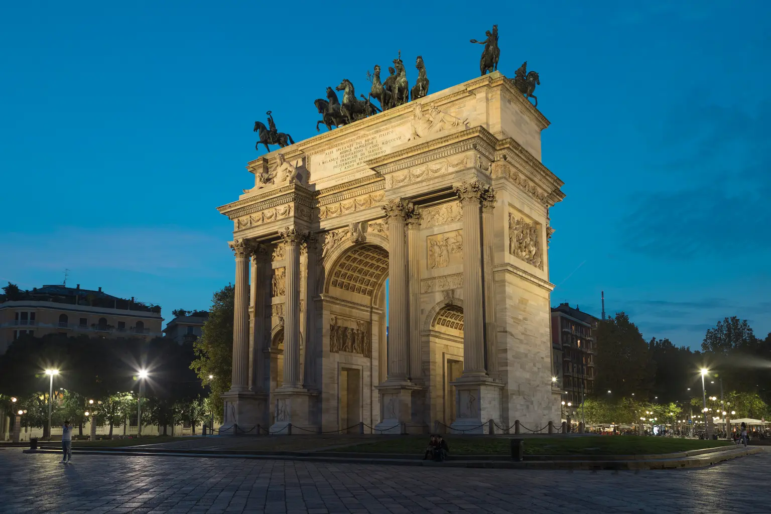 L'Arc de triomphe du Simplon est vu de côté au crépuscule, entouré d’arbres et des lumières de la ville, sous un ciel bleu profond en soirée.