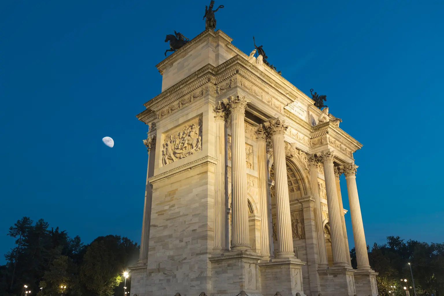 L'Arc de triomphe du Simplon est capturé sous un certain angle au crépuscule, avec ses détails architecturaux mis en valeur par l’éclairage et la lune brillant dans le ciel.