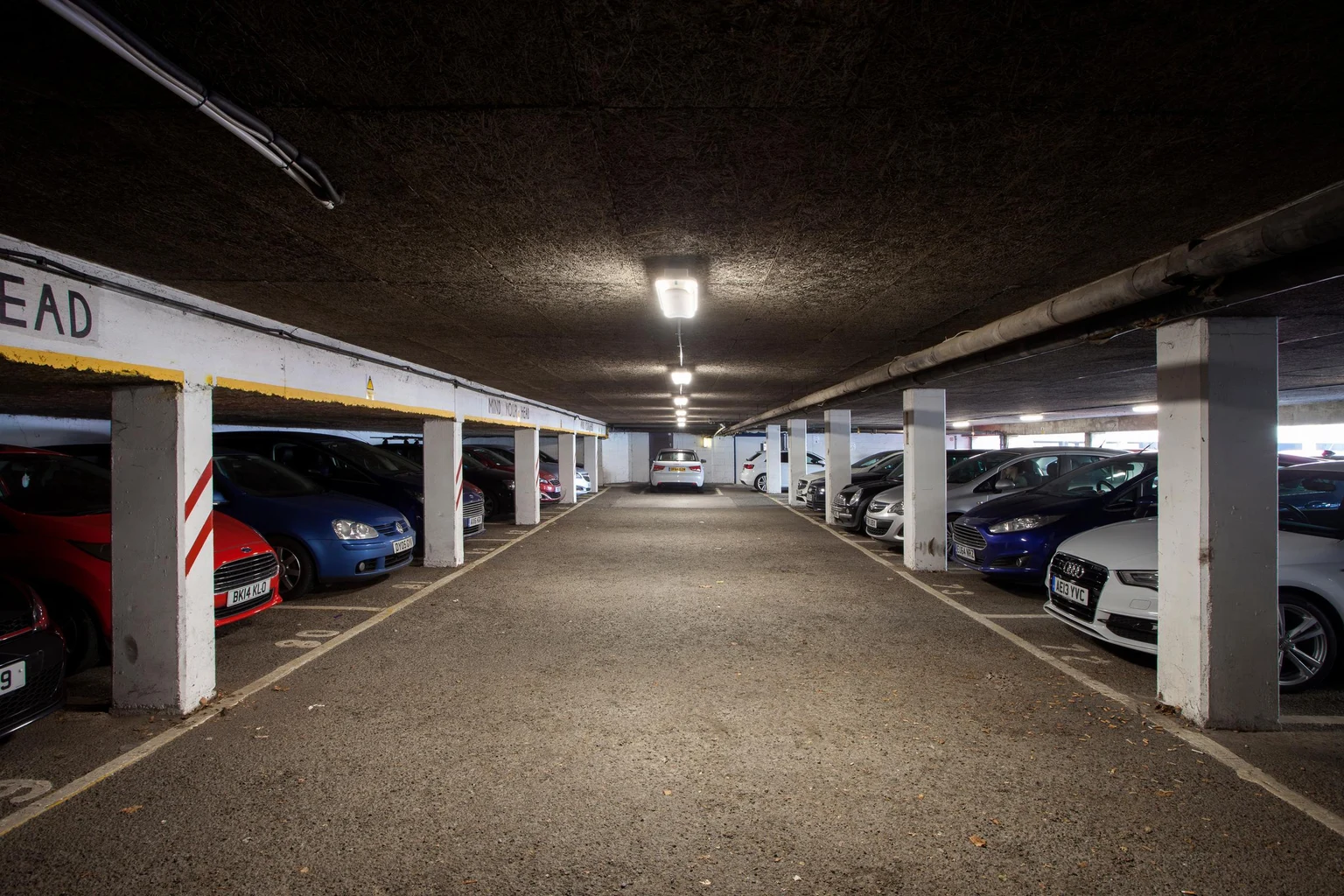 Un parcheggio coperto con file di auto parcheggiate su entrambi i lati e pilastri di cemento che sostengono il soffitto basso. Lo spazio è illuminato da luci fluorescenti a soffitto.