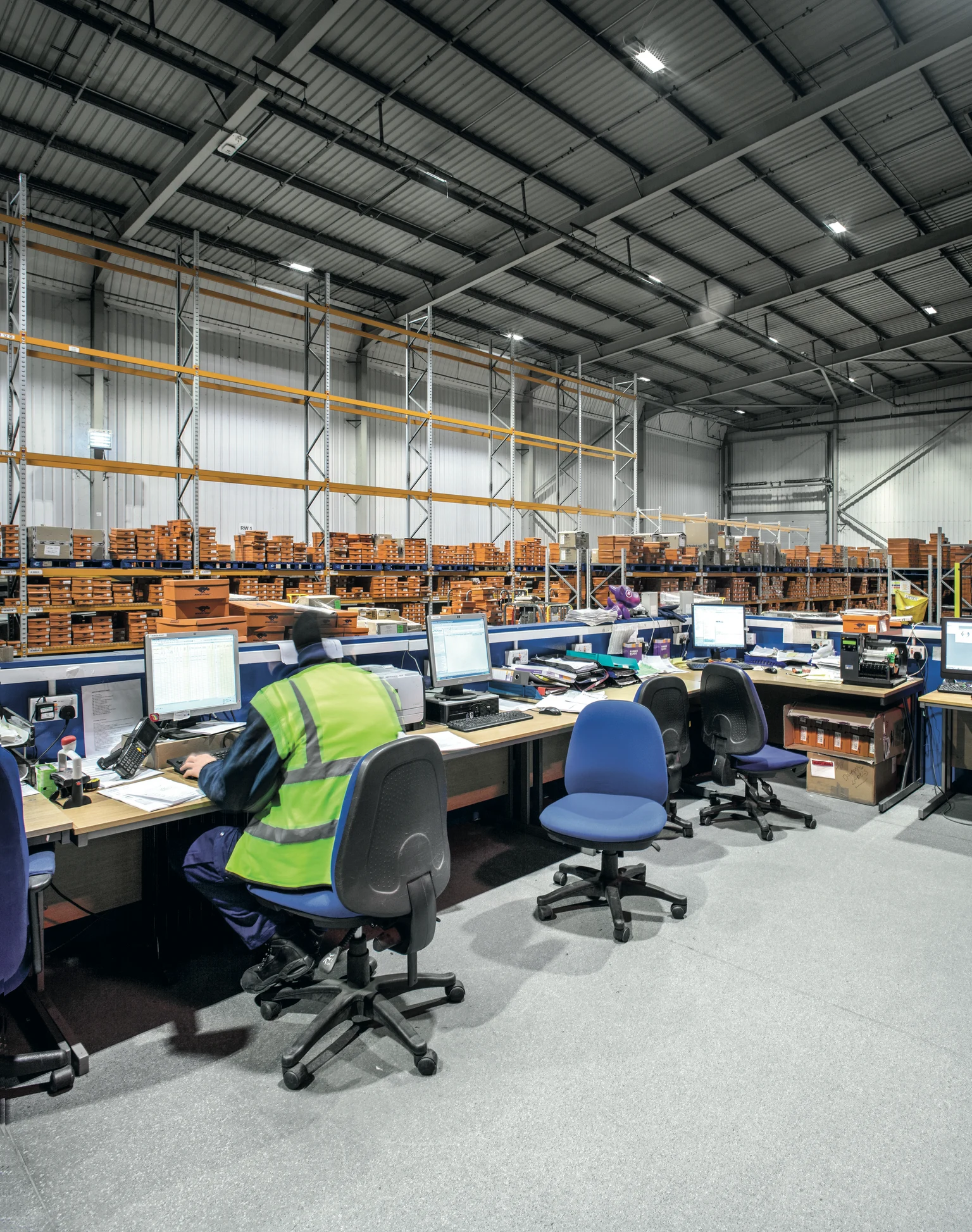Kantoorgedeelte in een magazijn, verschillende werkplekken met computers en een medewerker in een veiligheidsvest. De verlichting met aan het plafond gemonteerde ledlampen zorgt voor een goed verlichte en productieve werkruimte.