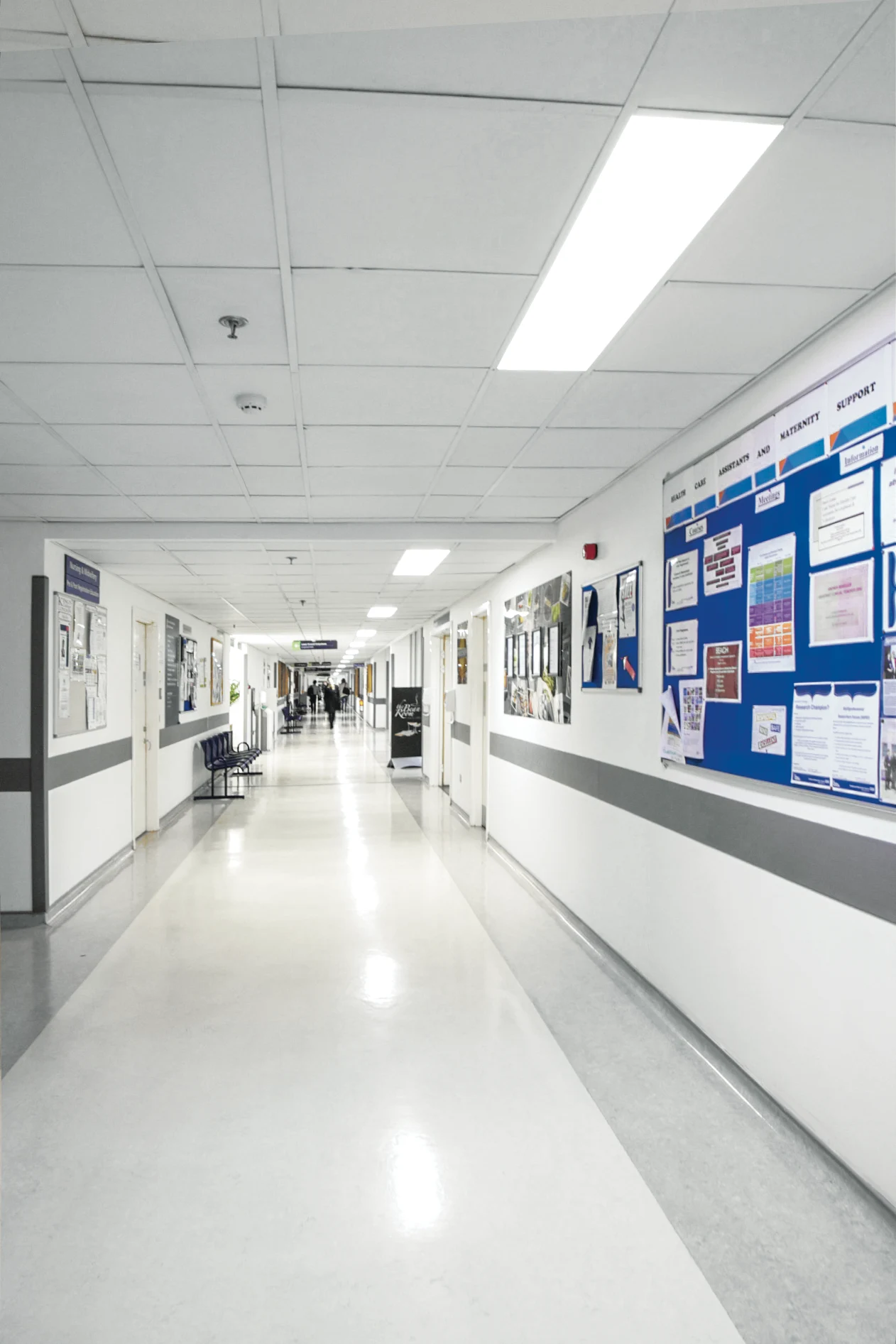 Lichte ziekenhuisgang met witte muren en grijze accenten. Aan prikborden hangen mededelingen. Verlicht door een ledpaneel.