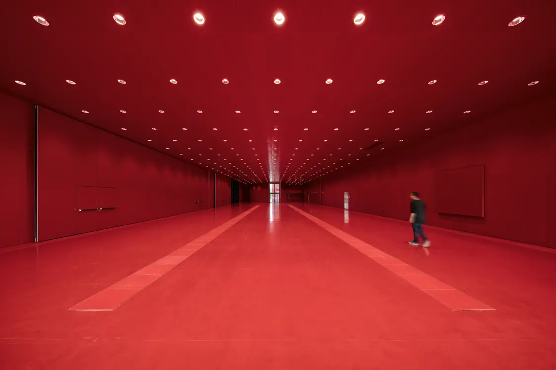 Un long espace rectangulaire entièrement habillé de rouge – sol, murs et plafond. La lumière est diffusée par des spots LED ronds, répartis de manière uniforme sur le plafond. Minimaliste et symétrique, le lieu dégage une esthétique futuriste, accentuée par une ligne centrale qui structure l’espace. Une personne le traverse, renforçant l’effet spectaculaire et futuriste de l’installation.