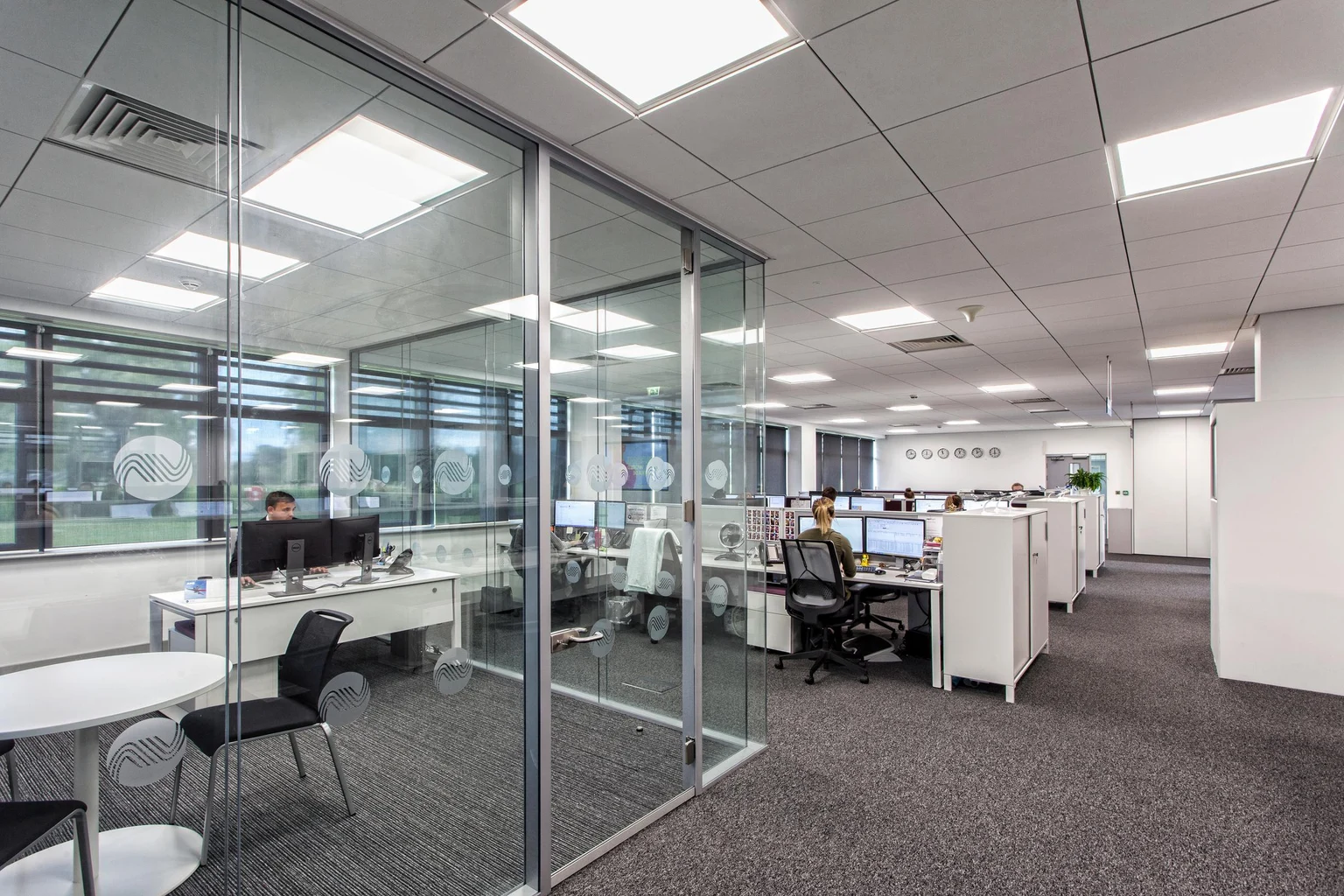 Een modern kantoor heeft glazen vergaderruimtes en rijen bureaus met computers, waar mensen aan het werk zijn. De omgeving is ruim, licht en goed georganiseerd.