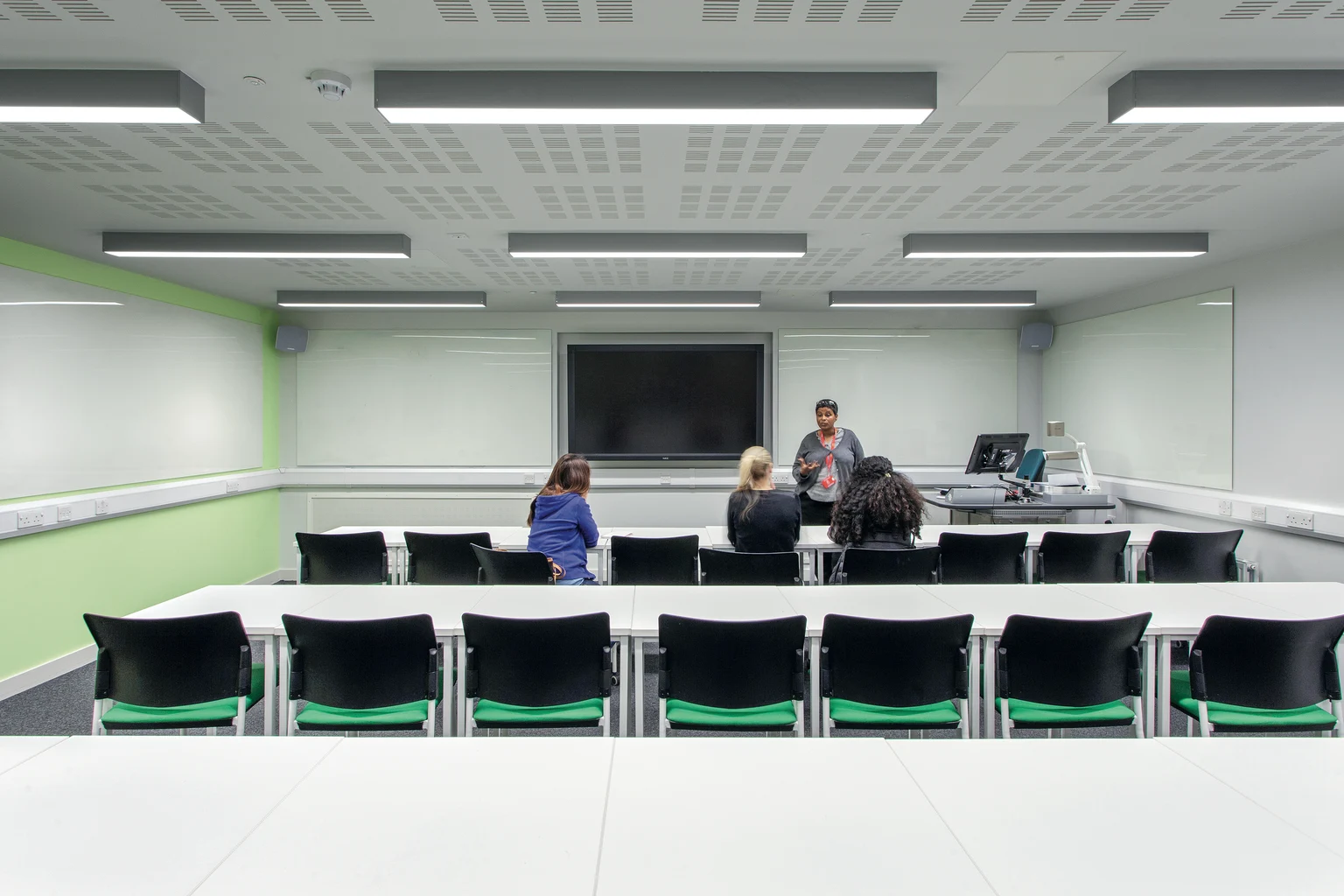 Een modern klaslokaal met rijen witte bureaus en zwarte stoelen, met vooraan een groot interactief scherm. De ruimte is helder verlicht met aan het plafond gemonteerde ledlampen en heeft muren met lichtgroene accenten.