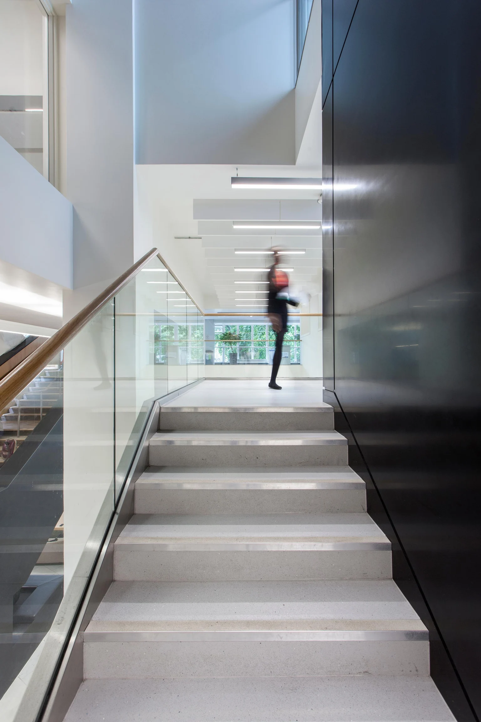 Een moderne trap met glazen balustrades en een houten leuning, die leidt naar een open, goed verlichte ruimte. Het ontwerp heeft strakke lijnen, witte muren en hangende plafondlampen voor een eigentijdse uitstraling.