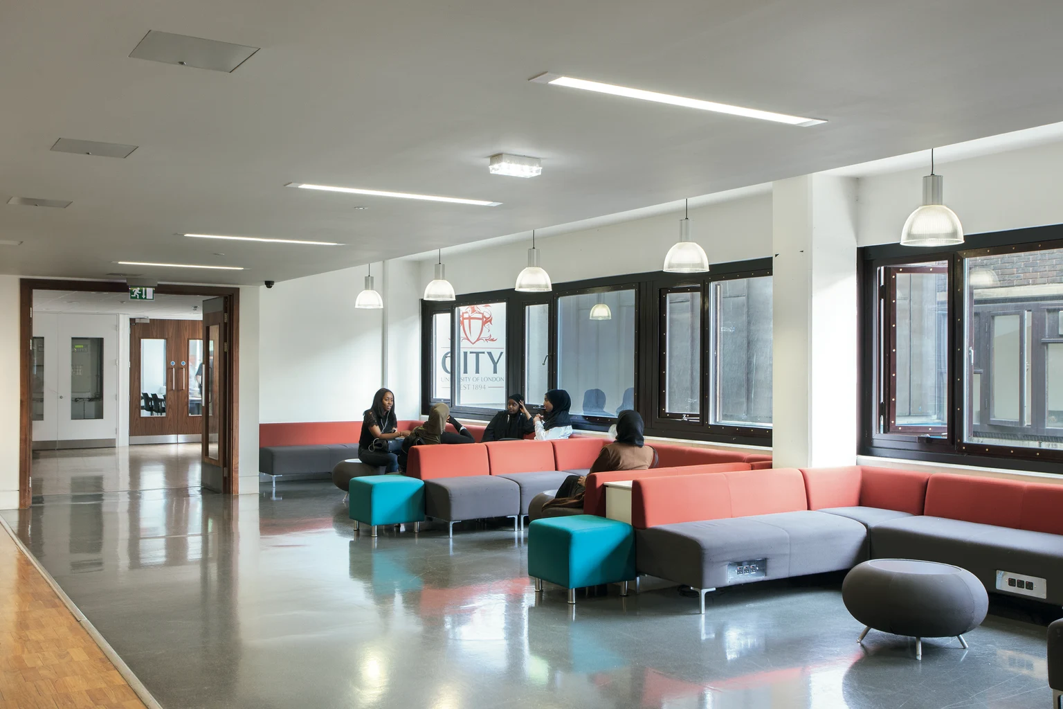 Een moderne ontspanningsruimte met kleurrijke zitplaatsen, waaronder rode en groenblauwe banken en grijze poefs. De ruimte is goed verlicht met hanglampen en inbouwspots, en heeft grote ramen voor een natuurlijke lichtinval.