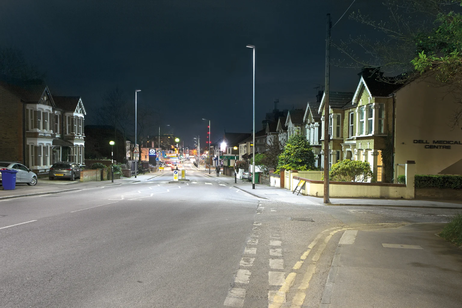 Een rustige straat in een woonwijk met aan weerszijden rijtjeshuizen, 's nachts verlicht door straatlantaarns. De straat lijkt leeg te zijn en de omgeving is rustig en vredig.