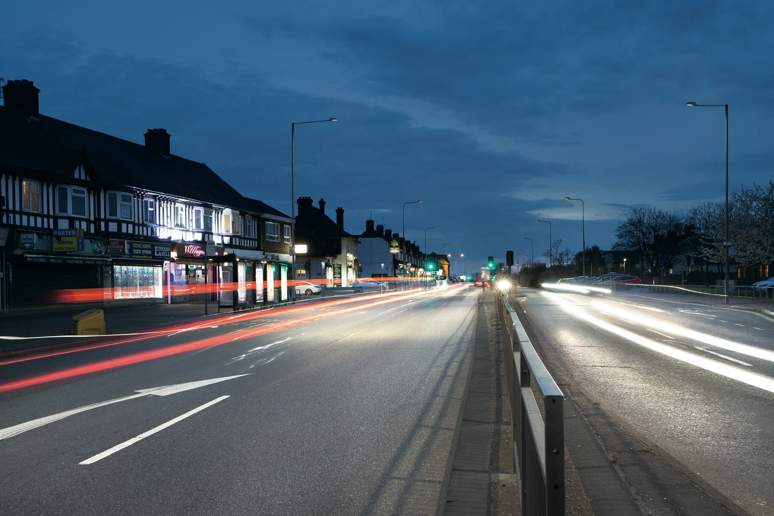 Een drukke straat bij schemering toont lichtsporen van passerende auto's, met erlangs gebouwen en straatverlichting. De lucht is donkerblauw. Het tafereel legt de beweging en energie van het avondverkeer vast.