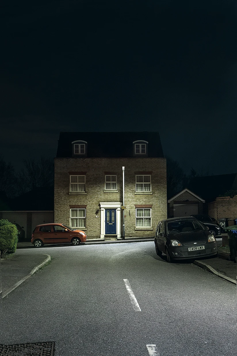 Aan het einde van een rustige straat staat een bakstenen huis, die ´s nachts wordt verlicht door een enkele straatlantaarn. Er staan twee geparkeerde auto's voor het huis en het tafereel is rustig en verlaten.