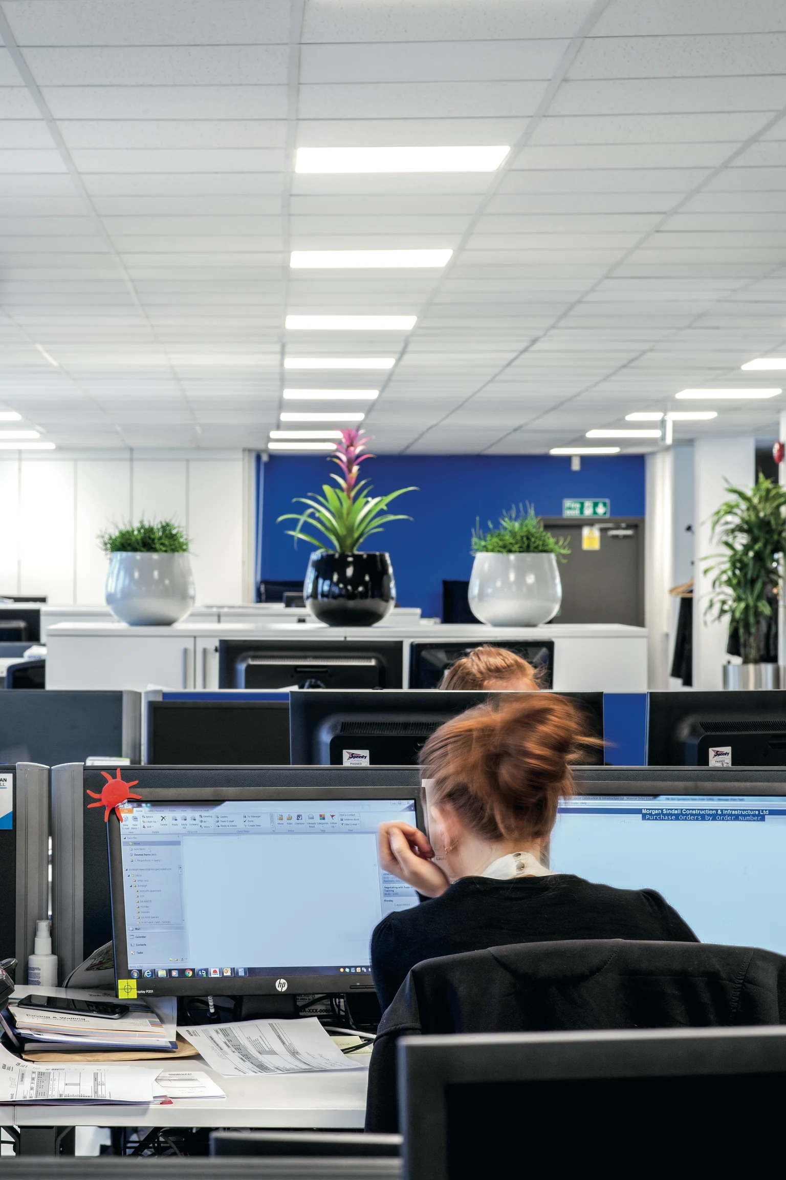 Un ufficio moderno presenta una sala riunioni con pareti di vetro accanto a postazioni di lavoro aperte con computer e sedie da ufficio. La disposizione è aperta e ariosa, con abbondante luce naturale e artificiale.