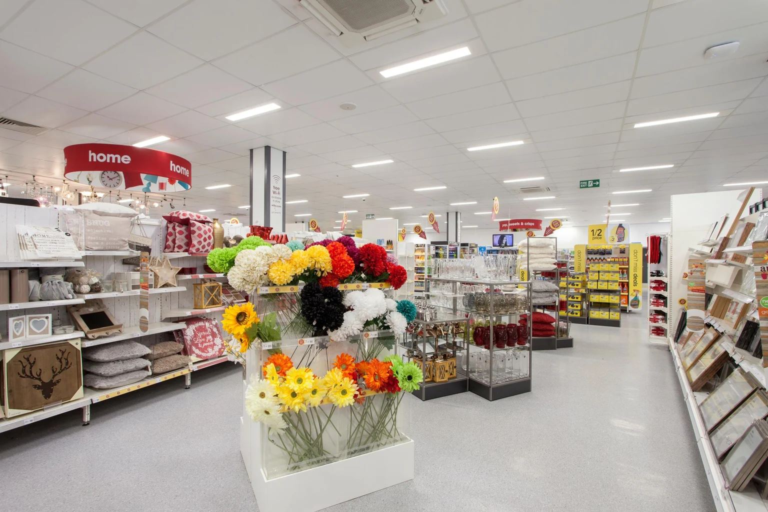 Un rayon d’articles ménagers dans un supermarché présentant des fleurs artificielles aux couleurs vives, des objets décoratifs et des étagères soigneusement disposées. La zone est bien éclairée avec un panneau « home » visible en arrière-plan.