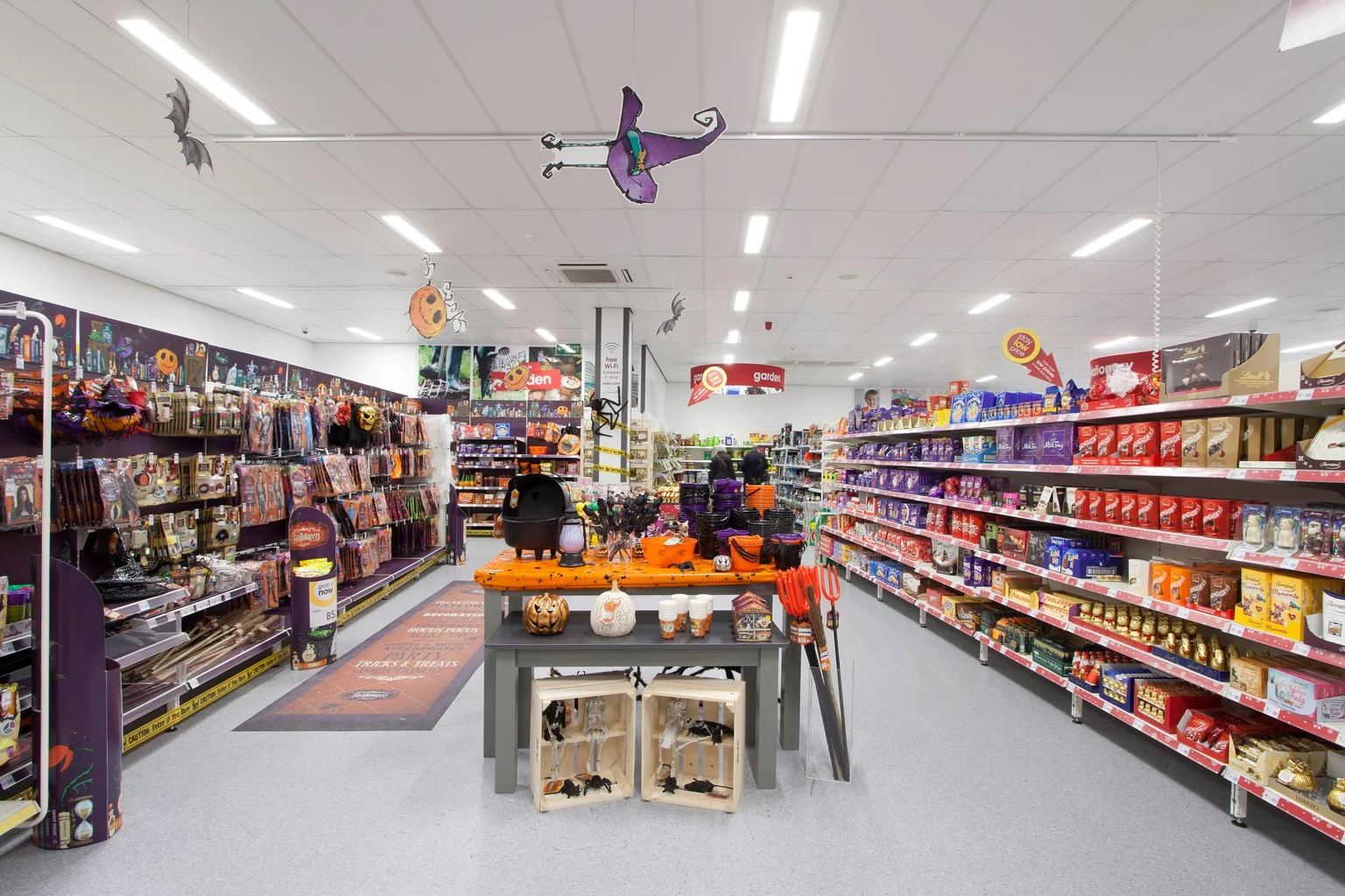 Une allée de supermarché festive décorée pour Halloween, avec des articles à thème tels que des citrouilles, des costumes et des décorations effrayantes. Les étagères sont garnies de produits emballés aux couleurs vives, et la table centrale expose des produits saisonniers.