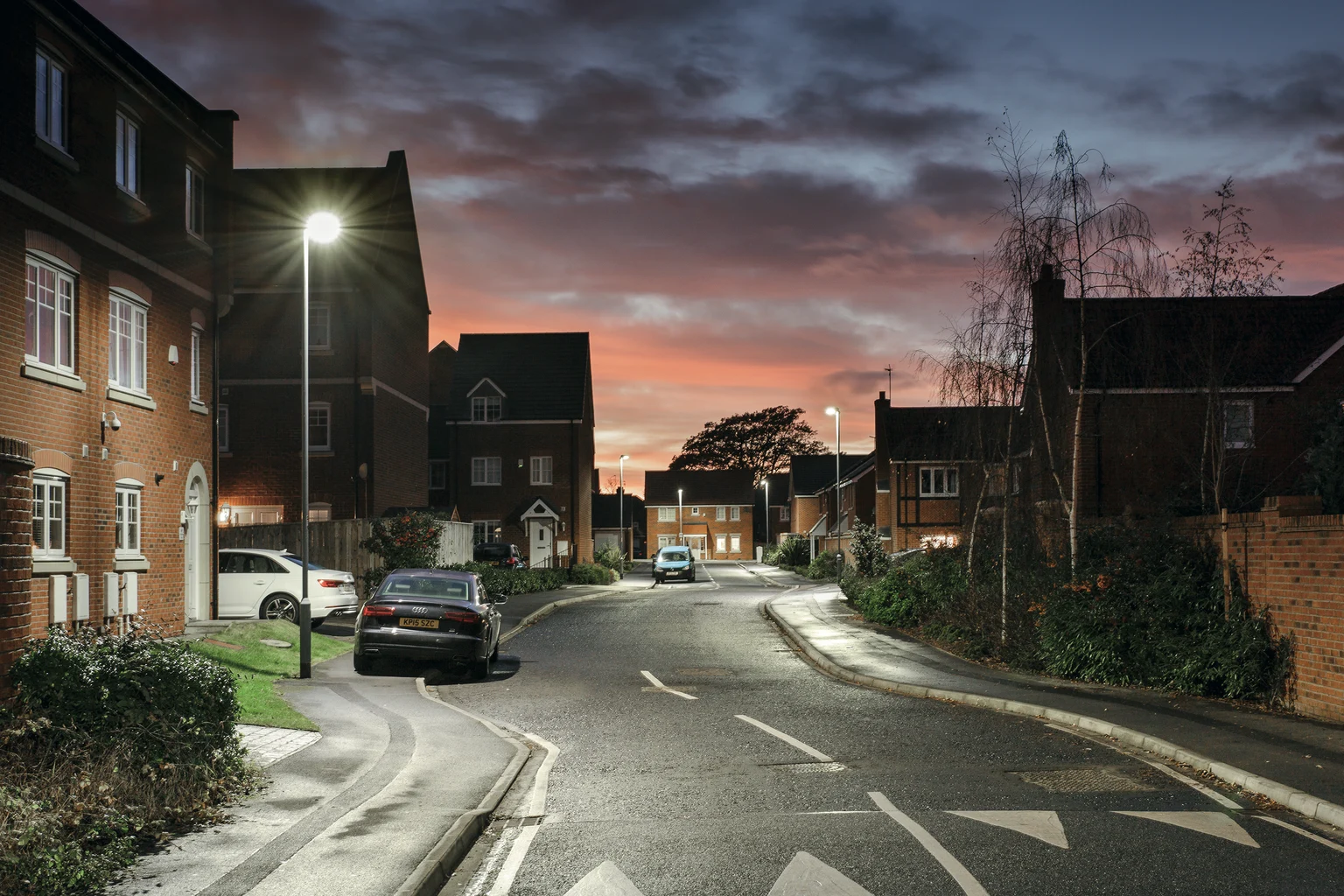 Een rustige straat in een woonwijk met geparkeerde auto's erlangs, bij schemering verlicht door straatlantaarns. De lucht straalt warme kleuren uit naarmate de avond nadert.