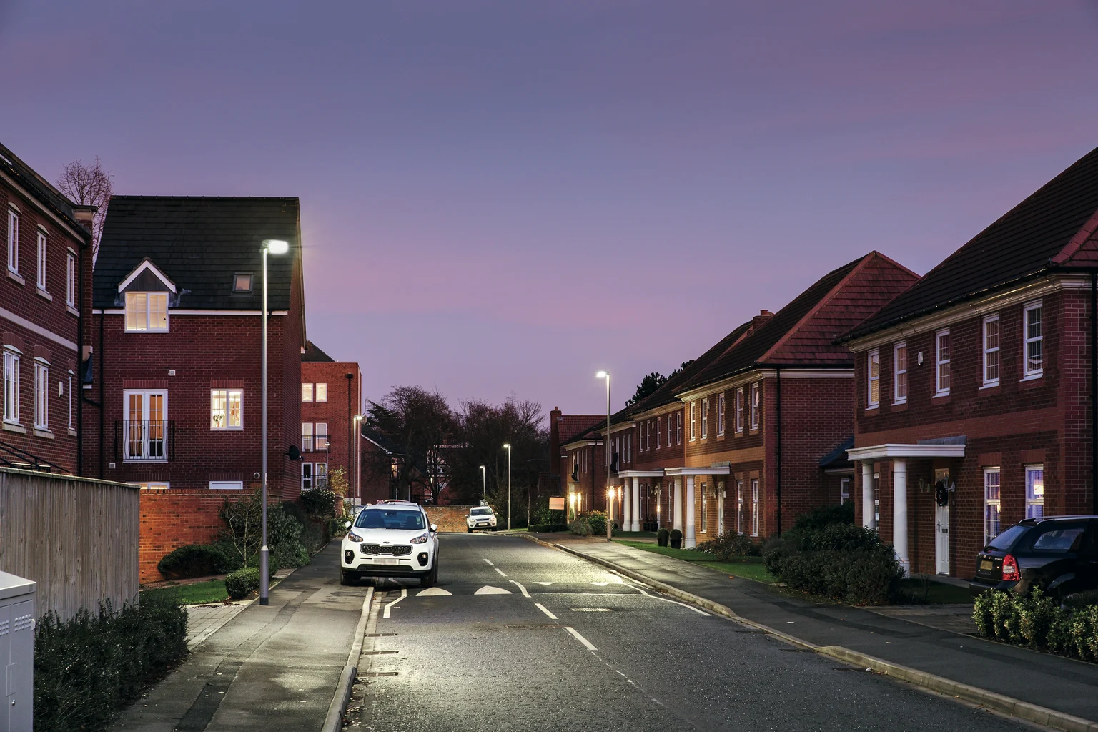 Een rustige straat in een woonwijk bij schemering, met moderne huizen en verlicht door straatlantaarns. Een witte auto staat geparkeerd aan de kant van de natte weg en weerspiegelt het zachte avondlicht.