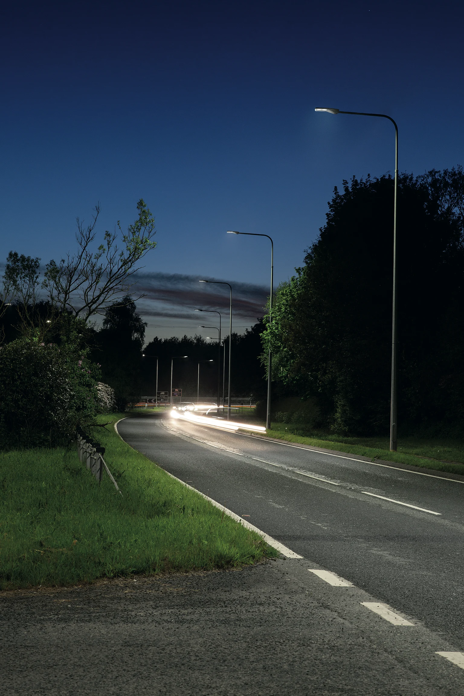 Een voorstedelijke weg met bocht bij schemering, verlicht door moderne straatverlichting en omgeven door bomen en groen. Lichtsporen van passerende auto's creëren een dynamisch effect onder een donkerblauwe avondlucht.