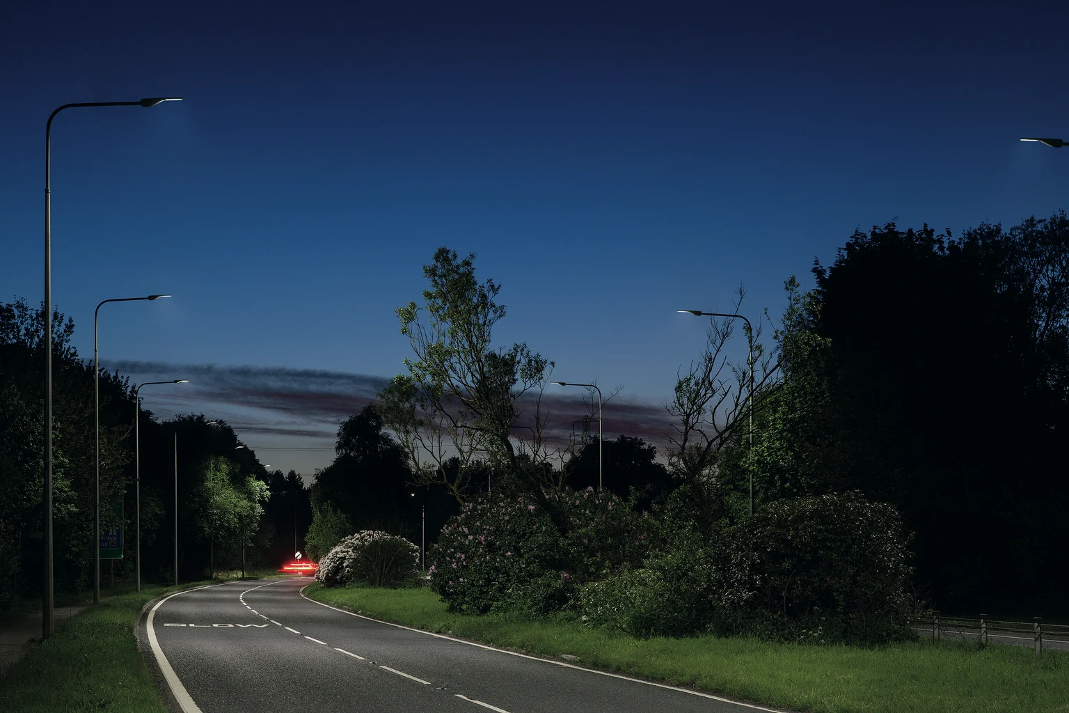 Een rustige voorstedelijke weg bij schemering, verlicht door moderne straatverlichting met bomen en struiken langs de zijkanten. De lucht vertoont een verloop van blauw en paars, en in de verte zijn de rode achterlichten van een auto zichtbaar.
