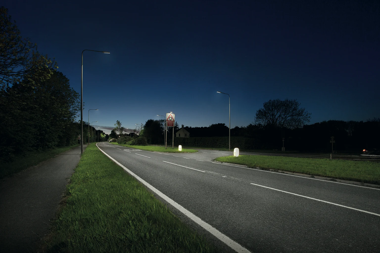 Een rustige voorstedelijke weg 's nachts, verlicht door moderne straatverlichting met grasvelden en bomen aan beide zijden. De lucht is donkerblauw en in de verte zijn huizen zichtbaar.
