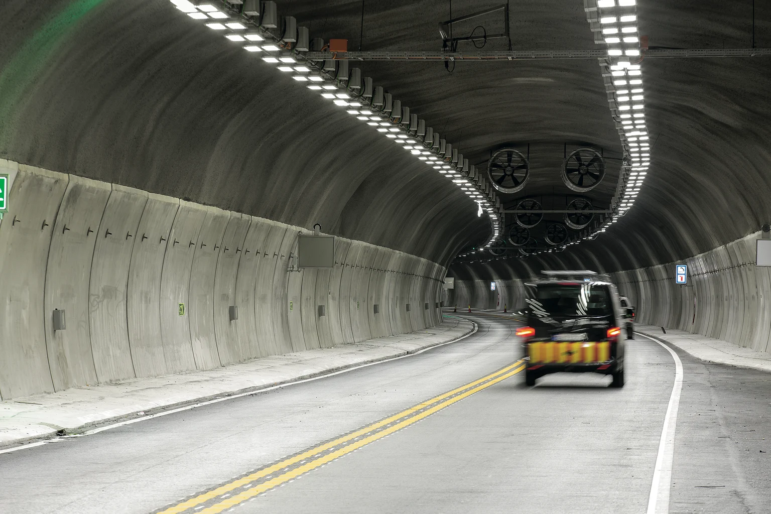 Un tunnel routier incurvé avec des murs en béton lisses et de puissants éclairages LED au plafond espacés uniformément. Un véhicule noir roule sur une route à deux voies marquée de lignes jaunes et blanches, tandis que de grands ventilateurs d’aération sont visibles à l’extrémité du tunnel.