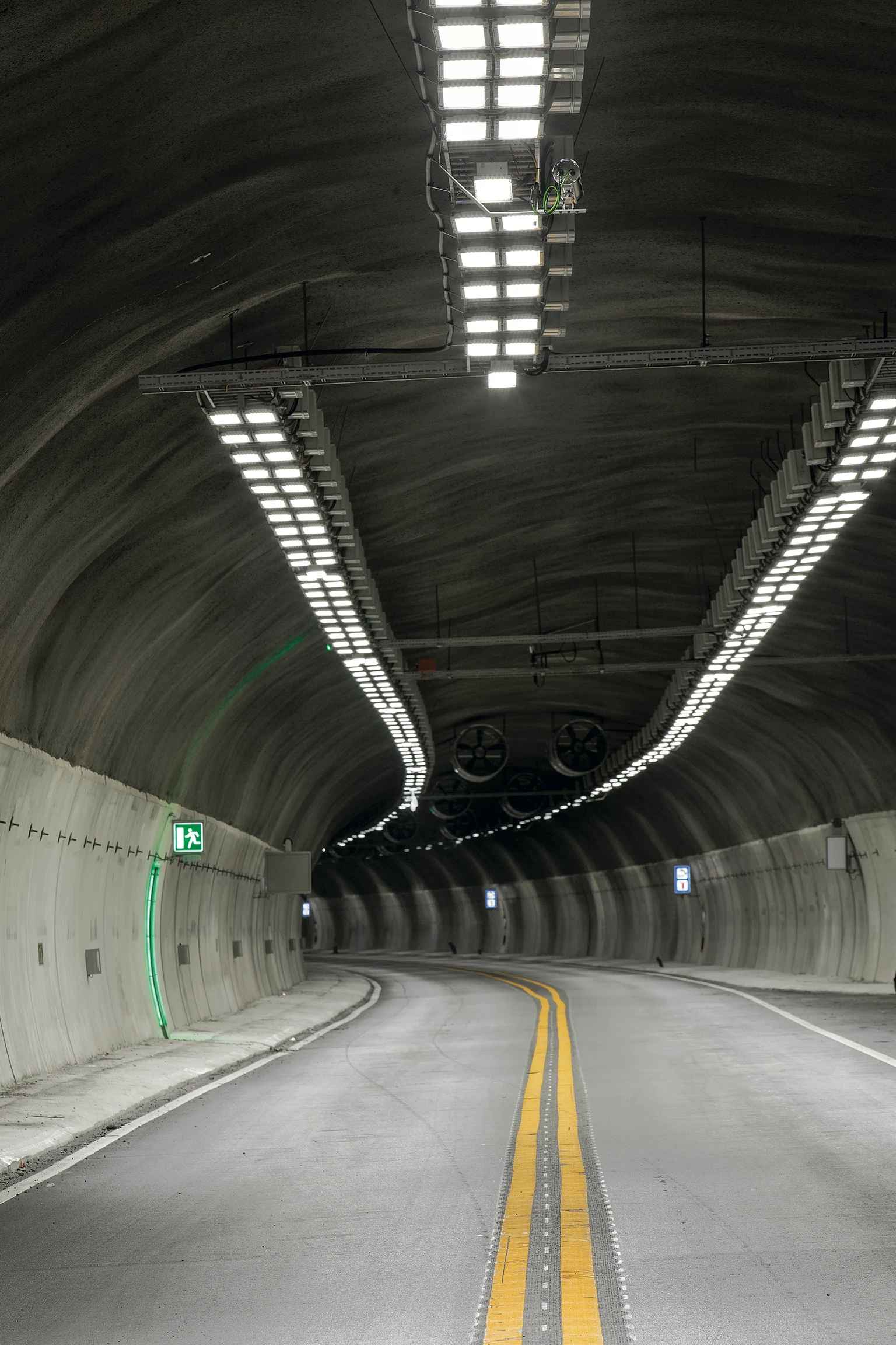 : un tunnel routier incurvé avec des murs en béton lisses, de puissants plafonniers LED et un panneau de sortie de secours vert lumineux à gauche. La route à deux voies est marquée de lignes jaunes et dispose d’un tracé ouvert pour un flux de circulation efficace.
