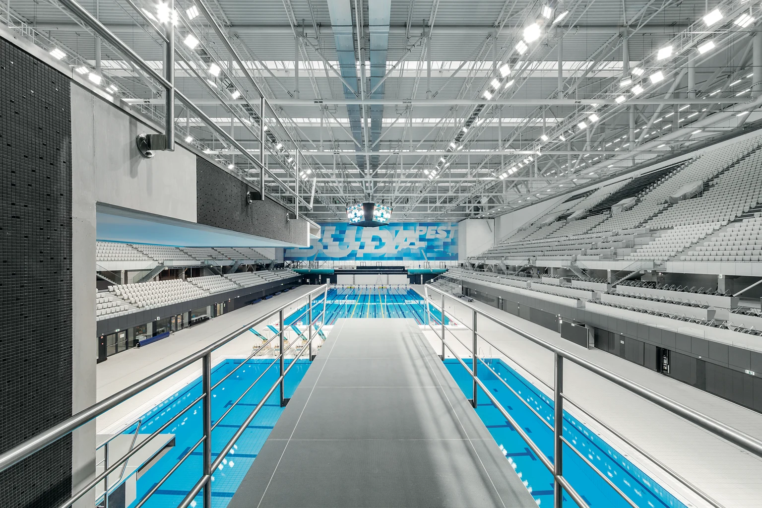 Modern binnenzwembad met meerdere banen, weergave vanaf een duikplatform. De hal is ruim en helder verlicht, met rijen zitplaatsen voor toeschouwers.