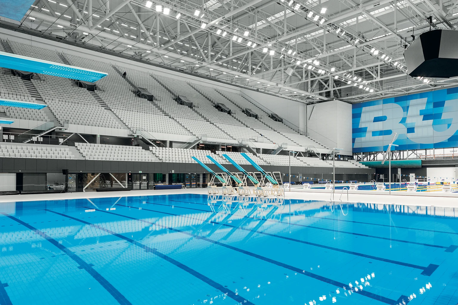 Modern binnenzwembad met meerdere banen, weergave vanaf een duikplatform. De hal is ruim en helder verlicht, met aan beide zijden rijen zitplaatsen voor toeschouwers.