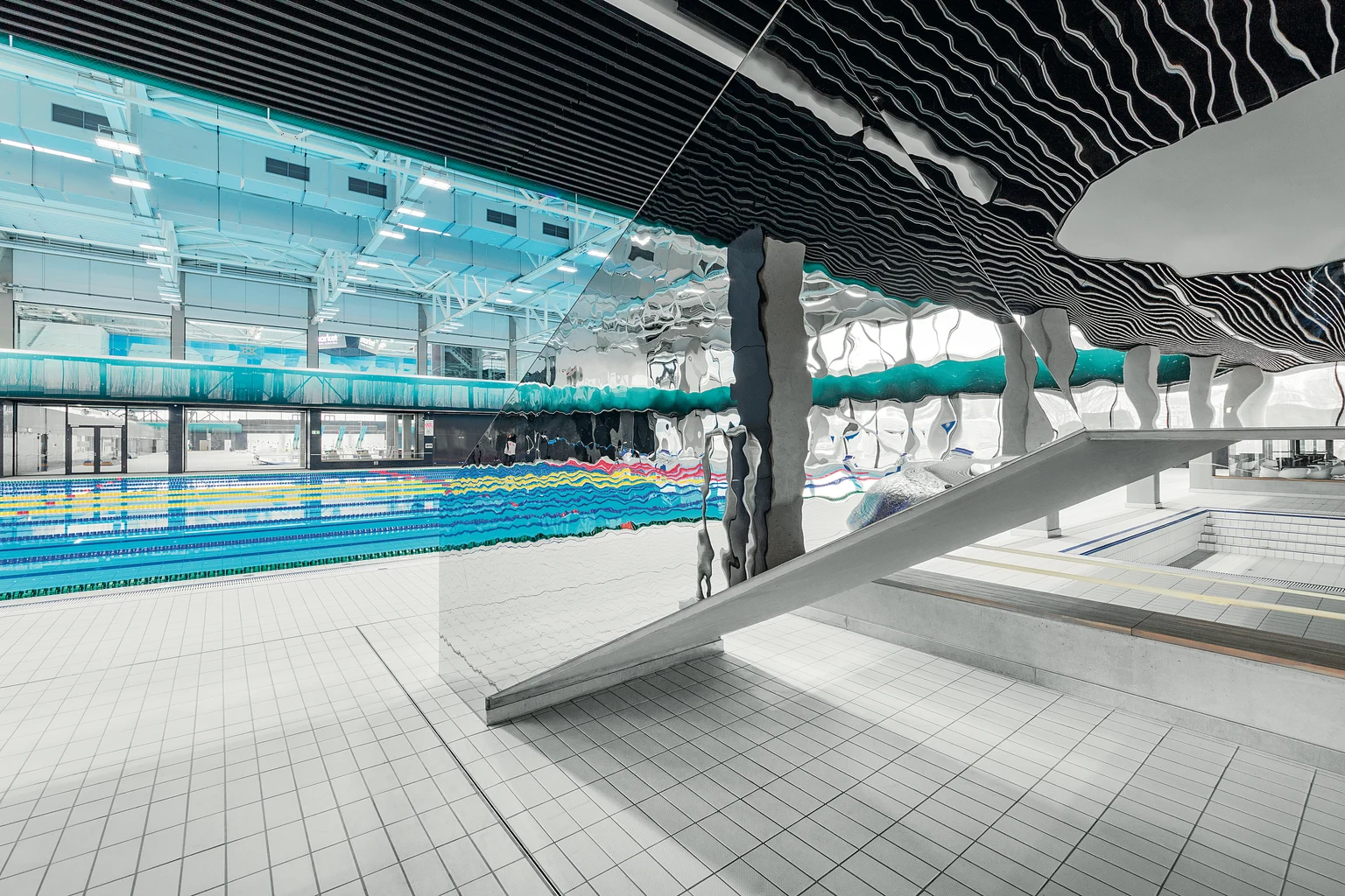Modern binnenzwembad met meerdere banen en een uniek, kunstzinnig plafondontwerp. De ruimte is ruim en gevuld met natuurlijk licht, met een glijbaan en zitplaatsen bij het zwembad.