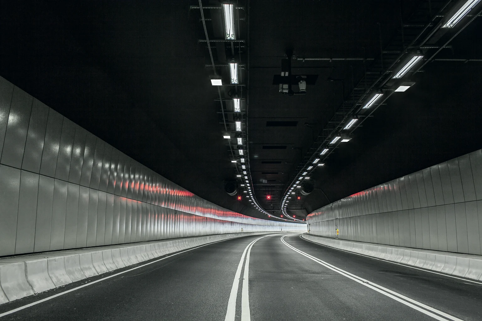 Een moderne, lege tunnel met heldere bovenlichten en witte rijstrookmarkeringen maakt een flauwe bocht naar links. De tunnelwanden zijn glad en reflecterend, wat een heldere en symmetrische uitstraling creëert.