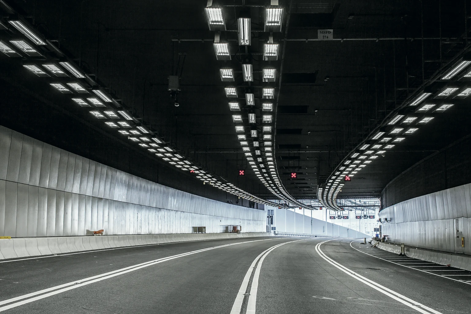 Een lege, goed verlichte tunnel met gladde betonnen wanden en heldere bovenlichten buigt flauw naar links. De weg is voorzien van heldere witte rijstrookmarkeringen en leidt naar de uitgang van de tunnel.