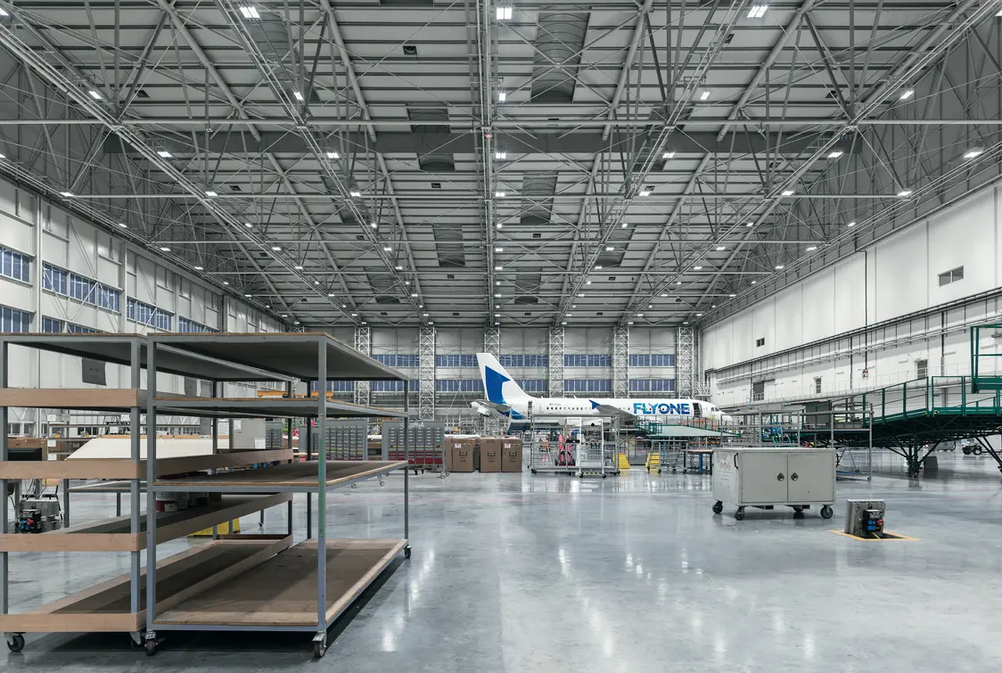 Große, moderne Flugzeugwartungshalle mit hoher Decke und viel Tageslicht. Im Hintergrund steht ein Passagierflugzeug, im Vordergrund befinden sich leere Regale und technisches Equipment.