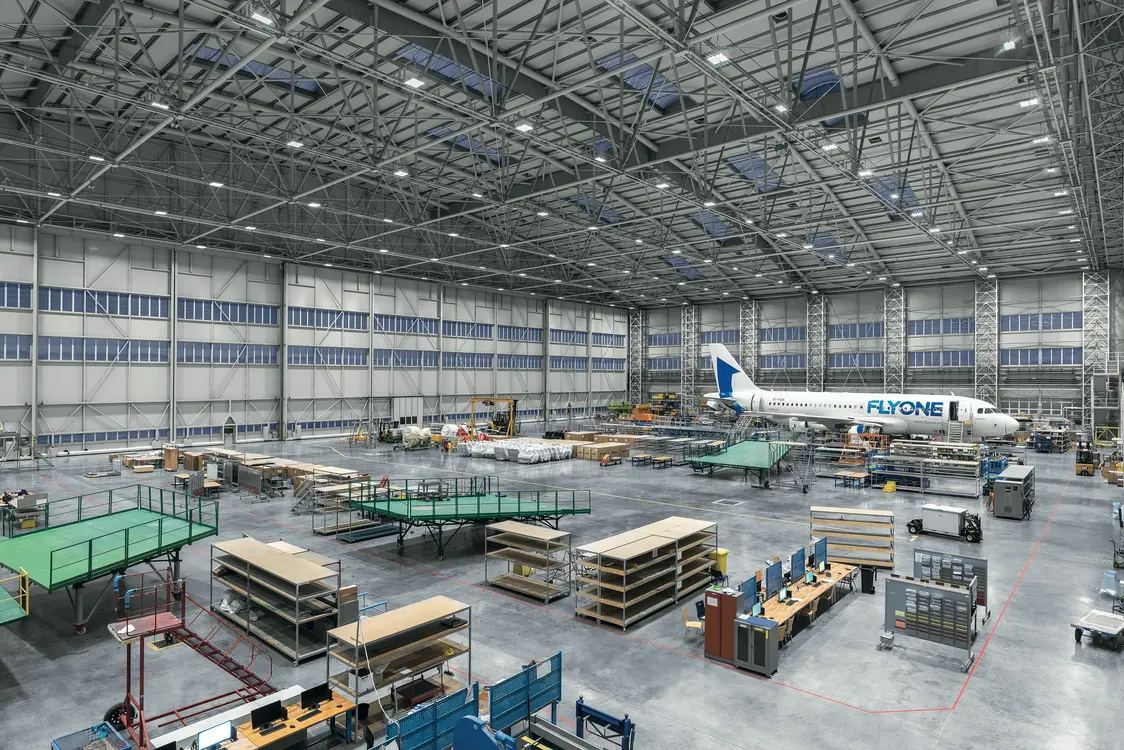 Große, moderne Flugzeugwartungshalle mit hoher Decke, vielen Arbeitsplätzen und einem Passagierflugzeug im Hintergrund. Die Halle ist hell beleuchtet und mit Werkbänken, Regalen und technischem Equipment ausgestattet.