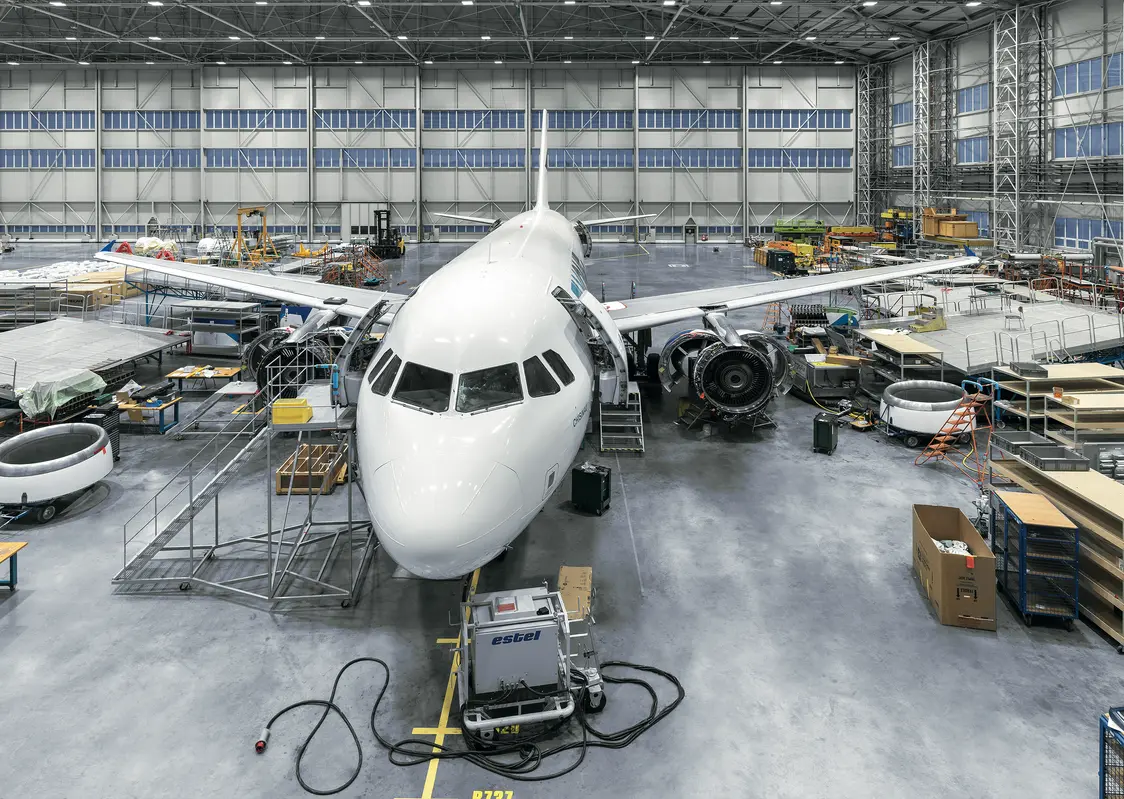 Großes Passagierflugzeug in einer hellen Wartungshalle mit hoher Decke und zahlreichen Arbeitsbereichen. Um das Flugzeug herum stehen Werkbänke, Wartungsgeräte und Materialkisten.