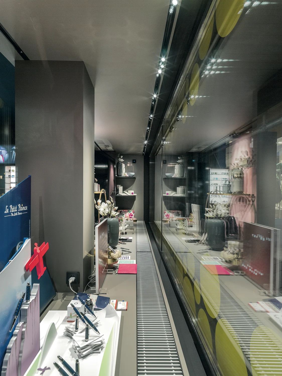 A narrow, modernly designed salesroom with illuminated shelves showcasing stylish accessories. A glass wall reflects the products and enhances the elegant atmosphere.