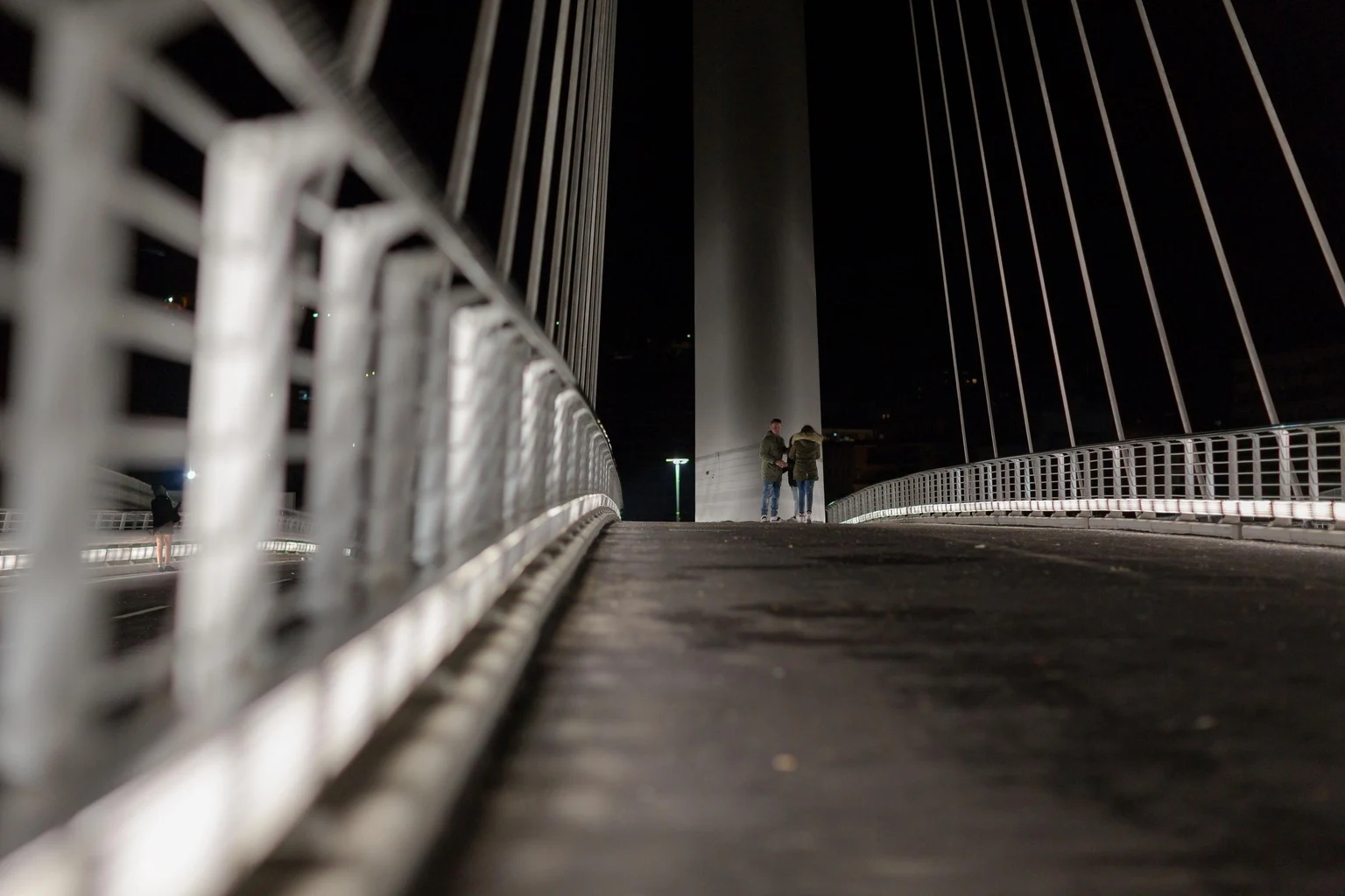 Eine moderne Schrägseilbrücke, bei Nacht fotografiert, mit zwei Personen, die in der Nähe des Mittelpfeilers stehen. Die Aufnahme aus niedriger Perspektive hebt Geländer und Seilkonstruktion hervor.